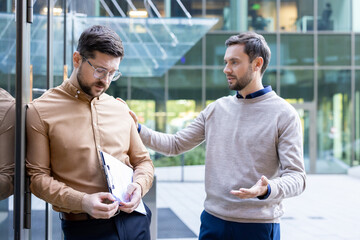 Two male colleagues standing outside an office building, one comforting the other who is looking down and holding documents, possibly after experiencing job stress or a difficult conversation