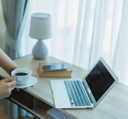 Fototapeta premium Woman taking a coffee break while working on a laptop at a modern wooden desk by a bright window, blending productivity and relaxed home office comfort during morning work