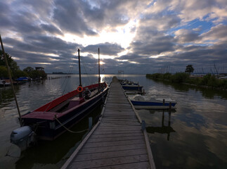 Obraz premium Wooden jetty with moored boats under a dramatic, cloudy sky at sunrise, Lake Dümmer in the morning in 49041 Damme, Germany