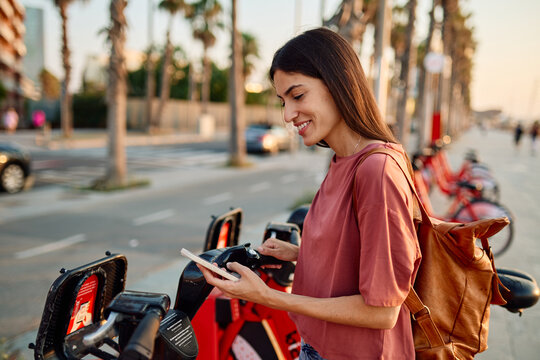 Smiling happy beautiful woman using mobile phone to unlock electric bicycle in city