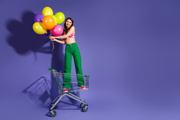 Cheerful young woman holding colorful balloons while standing on a shopping cart against a vibrant purple backdrop © deagreez