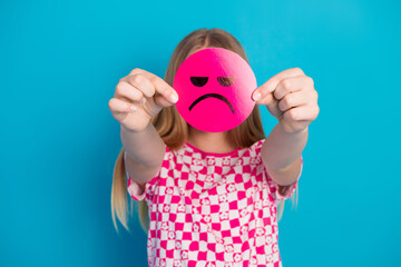 Young girl holding a pink unhappy smiley face against blue background - blonde teen serious expression