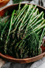 A bowl of green asparagus with a side of dill. The asparagus is fresh and healthy, and the dill adds a nice touch of flavor. The bowl is placed on a wooden table