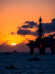 Naklejka premium offshore oil rig stands tall against a radiant sunset sky, its structural lights glowing as night approaches. Multiple boats circle in the distance, highlighting the ongoing operations in the open sea