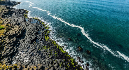 Aerial view of hexagonal basalt columns on the coast with turquoise ocean waves crashing against the shore, creating a white foamy line
