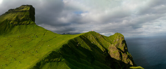 Scenic view of Kallur Lighthouse perched on a dramatic cliff edge on Kalsoy Island, Faroe Islands, surrounded by the North Atlantic Ocean and rugged coastal landscape under moody skies.