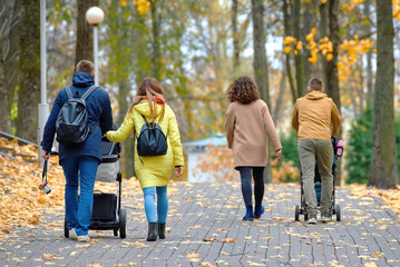 Families walking together in autumn park along paved pathway with colorful fallen leaves parents pushing strollers enjoying seasonal outdoor leisure and family bonding in fall environment