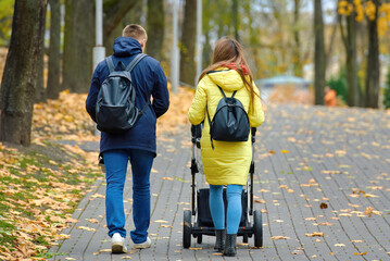 Autumn city scene with man walking in brown coat and stylish hat along urban sidewalk covered with yellow leaves, seasonal lifestyle solitude. Parents walk with their newborn through an autumn park.