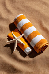 Orange striped towel and sunscreen with starfish on sandy beach background for summer vibes
