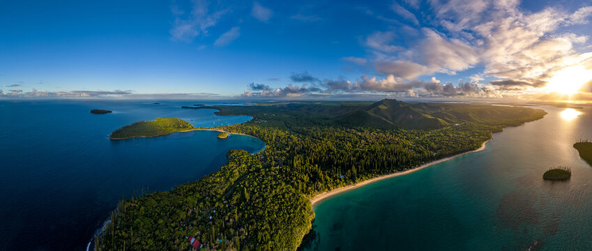Sweeping aerial panorama of Kuto Bay on the Isle of Pines, New Caledonia, during a vibrant sunrise. The lush green island is surrounded by calm, turquoise water under a beautiful sky