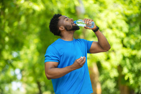 Young man drinking water from a plastic bottle while taking a break during outdoor workout. Athlete in blue t-shirt staying hydrated after running in a sunny park.