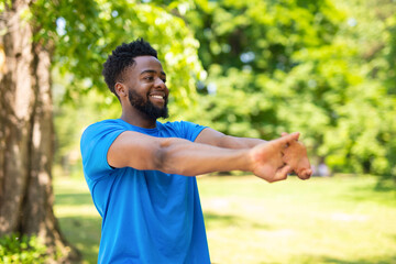 Smiling young man stretching arms outdoors in park, preparing for workout in blue sportswear.
