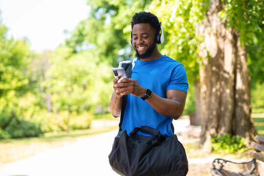 Happy young man in blue sportswear using smartphone and headphones in park after workout.
