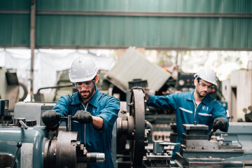 Two factory workers are collaborating on a large piece of industrial machinery. Both men are wearing blue jumpsuits, black work gloves, and white hard hats, with also wearing safety glasses.