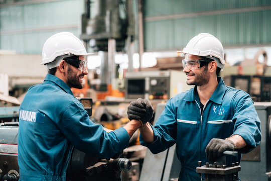 Two male factory workers, wearing identical blue jumpsuits, white hard hats, and safety glasses, are giving each other a fist bump.smiles on their faces and are looking at each other teamwork