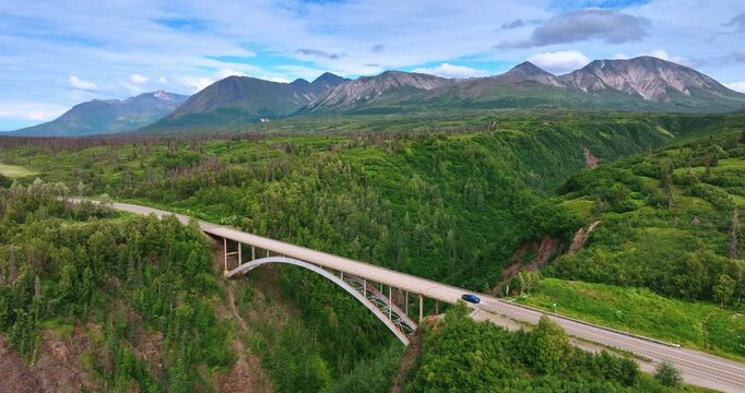 Blue car crosses the Hurricane Gulch Bridge in Alaska, USA. Beautiful green landscape and mountain range at backdrop.