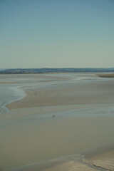 Panoramic view of the bay at Mont Saint-Michel, France. People walking on the sand during low tide with a small island in the distance, under clear sky. Peaceful coastal and travel scenery