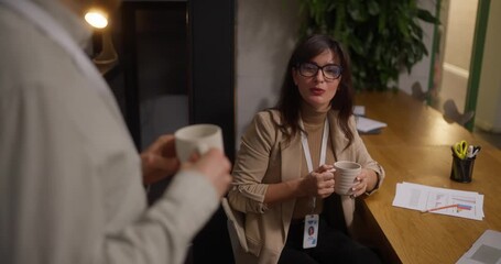 Over the shoulder Girl in business clothes holds mug of hot drink in her hands and chatting with her colleague while sitting at a table in a working setting During lunch - Powered by Adobe