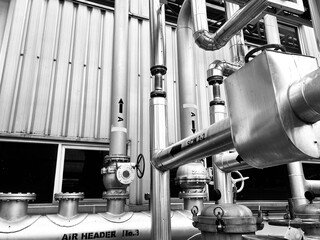 black-and-white image of complex industrial piping and machinery. The detailed composition of valves, tubes, and a corrugated metal wall highlights the intricate and functional aesthetics