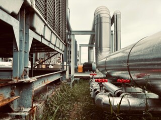 Large industrial pipes and a cooling tower at a factory or power plant, with a focus on the heavy-duty engineering and rugged outdoor setting. The image highlights the contrast between the worn metal 
