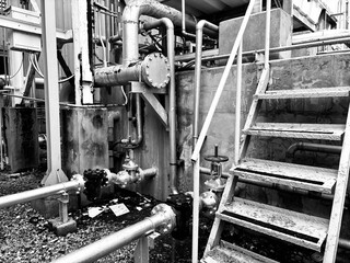 A black and white image of industrial machinery, pipes, and a metal staircase in a gritty factory environment. The photo captures the worn textures and complex layers of an aging facility, conveying 