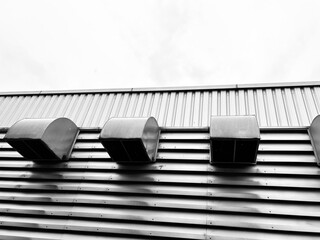 A close-up, low-angle shot of a building's corrugated metal wall, highlighting a series of industrial vents. The black and white photography emphasizes texture and modern, minimalist architecture.