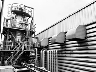 A dramatic black and white image of industrial tanks and pipes at a modern factory or chemical plant. The composition highlights the clean lines of the corrugated metal building and the complex arrang
