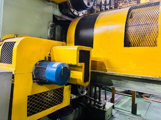 A vivid close-up of heavy industrial machinery in a factory, dominated by bright yellow metal casing and a contrasting blue electric motor. The image highlights the intricate details of the machine,