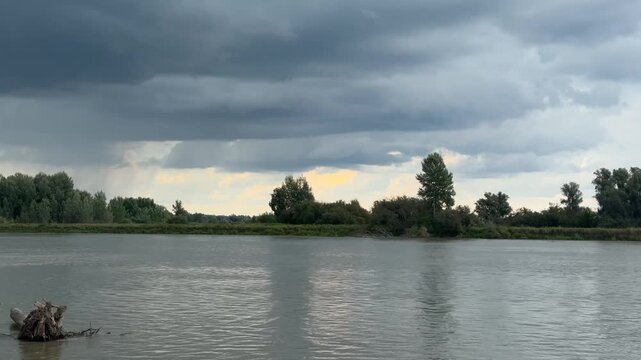 Wide river landscape on a cloudy day with a weathered driftwood in the foreground. Gentle breeze ripples the water. Color palette from dark gray to soft sky blue with bright breaks in rain clouds. Dis