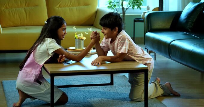 Indian small siblings playing arm wrestling panja game together at home indoors, Asian young kids siblings enjoying fun playful childhood activity in living room, family bonding, lifestyle, happiness