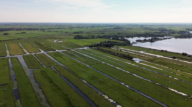 Aerial view of polder farmland with long green strips divided by water channels. A lake and scattered buildings in the background highlight effective land and water management in a rural setting.