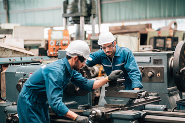 Two factory workers are collaborating on a large piece of industrial machinery. Both men are wearing blue jumpsuits, black work gloves, and white hard hats, with also wearing safety glasses.