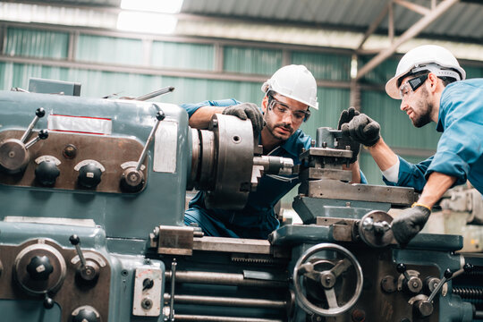 Two factory workers are collaborating on a large piece of industrial machinery. Both men are wearing blue jumpsuits, black work gloves, and white hard hats, with also wearing safety glasses.