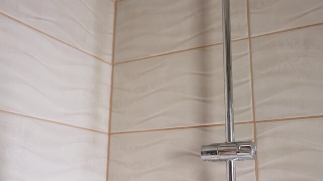 High angle tiled bathroom shows chrome shower on holder as female hand reaches, grips handle, lifts head from mount for washing, light glints on rippled tiles, calm interior, hygiene routine