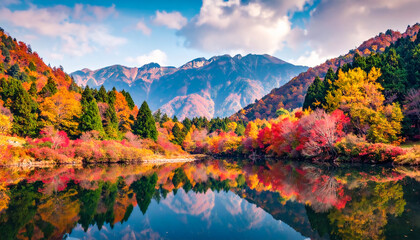 Obraz premium Stunning autumn foliage reflects in serene lake beneath dramatic mountains on a peaceful sunny day