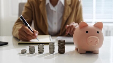 Business person writing financial notes with stacked coins and piggy bank, symbolizing savings, money management, and investment planning. - Powered by Adobe