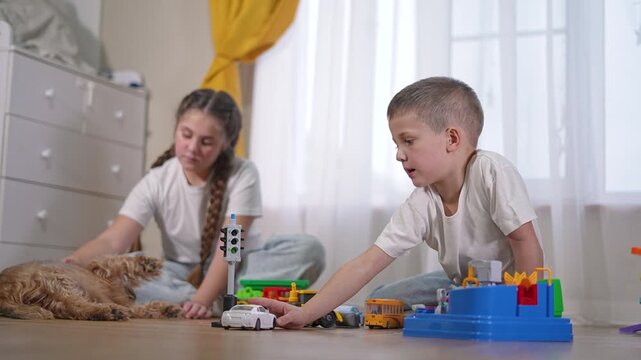 Boy lifts plastic traffic light over toy cars and trucks. Girl nearby strokes puppy lying on floor. Child explores objects in creative play. Boy, toy, child, puppy create playful composition.