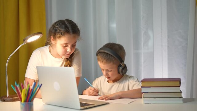 Girl sits beside boy with headphones. Both students look at laptop while discussing task. Child listens and keeps writing. Girl explains studying. Writing focus continues with laptop and headphones. - Powered by Adobe