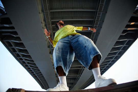 Man standing under bridge, arms outstretched, wearing oversized clothing, looking upward, low angle view emphasizing urban environment, representing Gen Z street culture