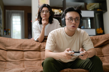 A man wearing a beige shirt sits on a brown couch, focused on a game controller with over-ear headphones, while a woman watches from behind