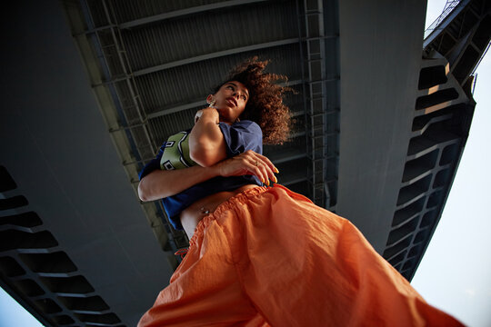 Young woman standing under bridge, looking away from camera, holding arm, curly hair blowing, embodying Gen Z style and attitude, urban setting, low angle perspective