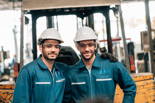 Two factory workers are standing together, with a large piece of yellow machinery, likely a forklift, visible behind them. They are both wearing blue jumpsuits, white hard hats, safety glasses