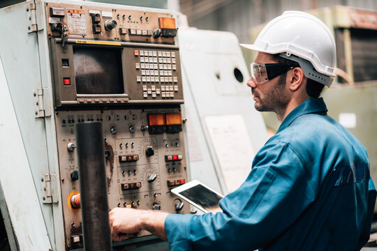 A young male factory worker, wearing a blue jumpsuit, a white hard hat, and safety glasses, is standing in front of a complex piece of industrial machinery. He is holding and looking down at a tablet