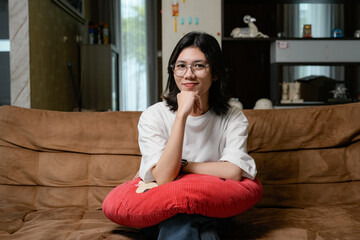 Obraz premium A relaxed woman in a white shirt sits on a brown couch, hugging a red pillow in a cozy living room