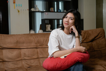Obraz premium A relaxed woman in a white shirt sits on a brown couch, hugging a red pillow in a cozy living room