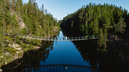 Visitors walk across a hanging suspension bridge over the serene Canyon Lake Julma Olkky, surrounded by lush forests in Hossa National Park in Finland