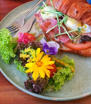 Croissant sandwich with ham, poached egg, tomato, and salad with edible flowers, served on a ceramic plate.
