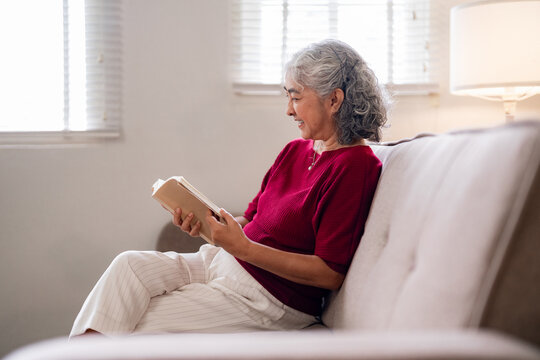 Asian older woman reading book happily living room peaceful domestic environment enjoying leisure time comfortable home senior female wellness