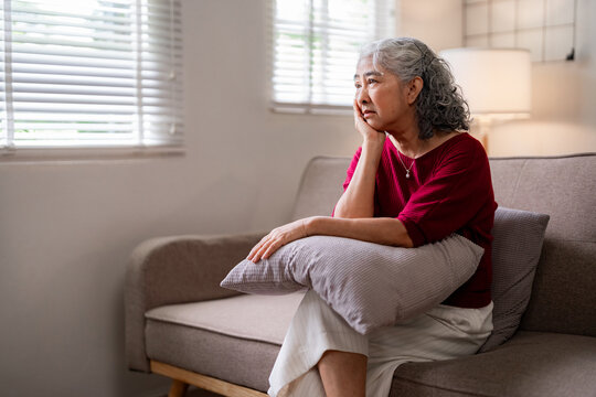 Older Asian woman sits quietly on home sofa contemplating window view Pensive senior female solitude in cozy living room domestic setting Mental wellness concept - Powered by Adobe