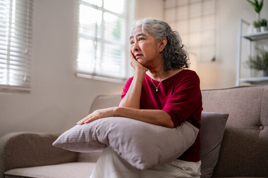 Older Asian woman deep thought home living room sofa contemplating life moments experiencing quiet reflection loneliness mental health
